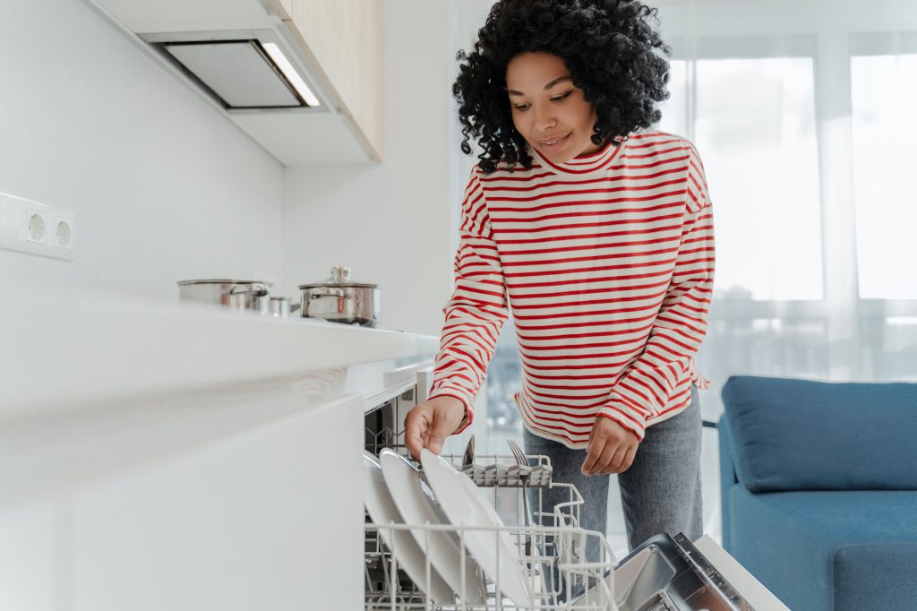 Beautiful African American woman loading dishwasher with dishes in kitchen at home