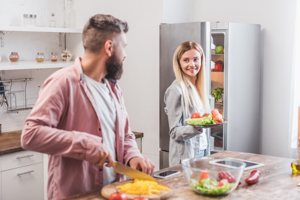 Husband cutting vegetables and wife standing near refrigerator