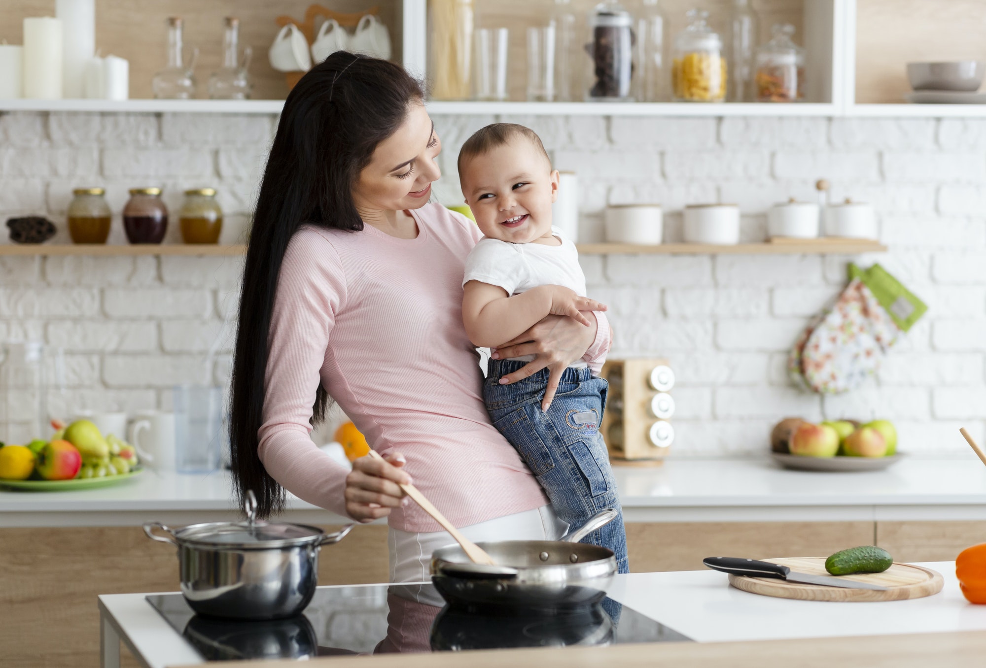 Millennial woman laughing with baby son, preparing food at kitchen