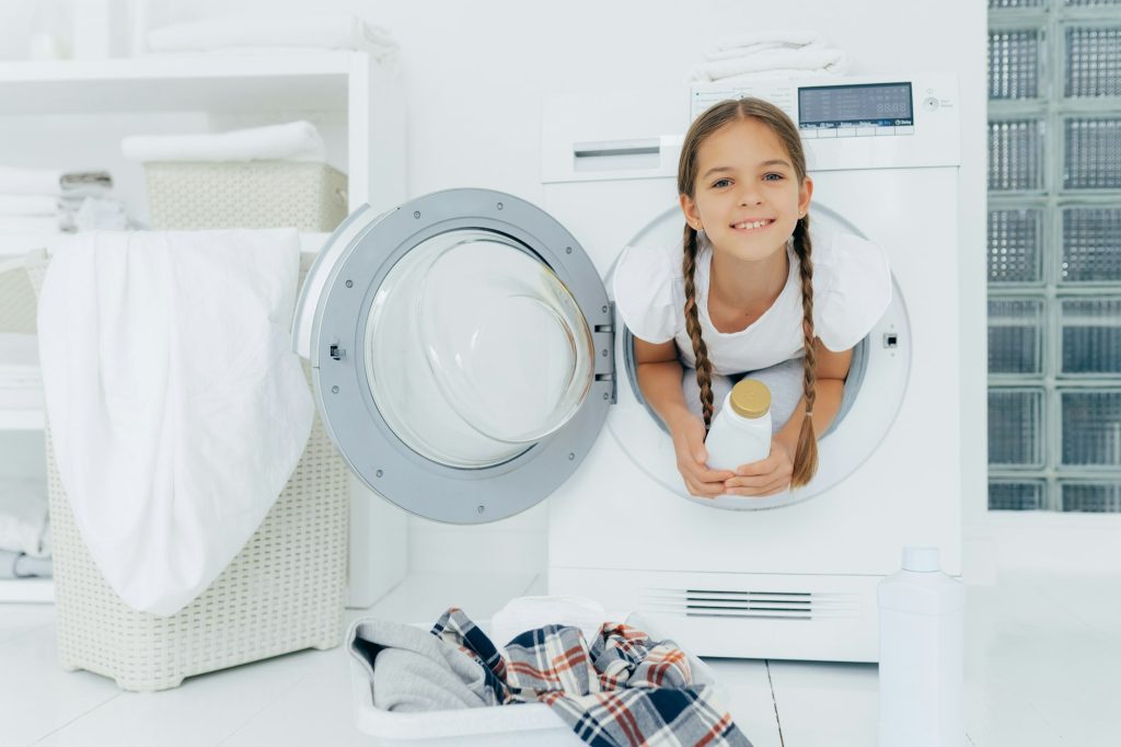 Smiling female child with glad expression, poses in washing machine, holds detergent