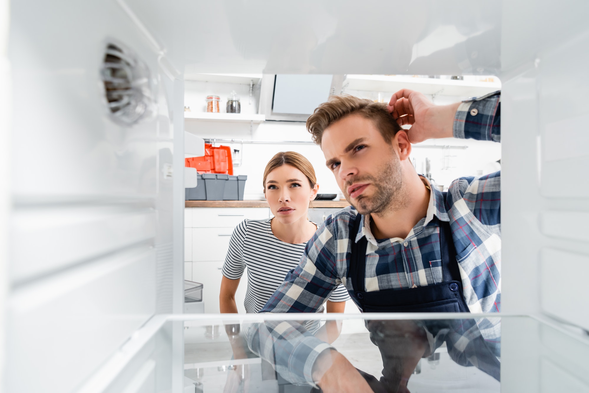 thoughtful handyman and young woman looking at freezer on blurred foreground in kitchen