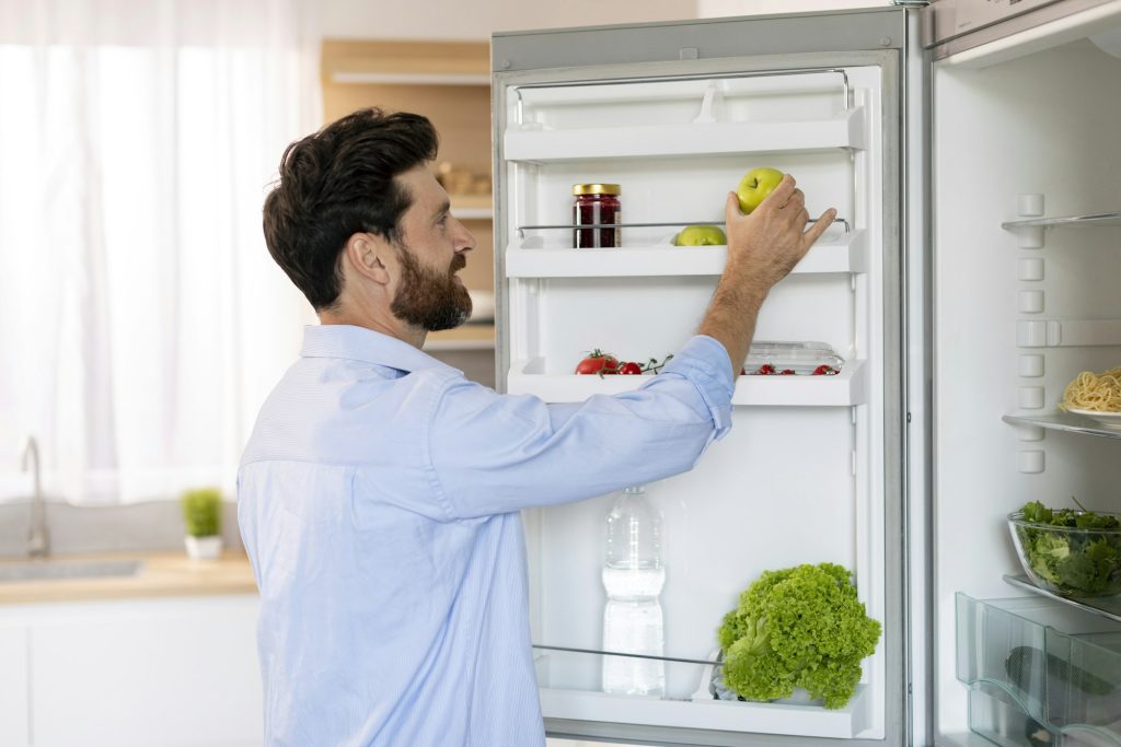 Happy hungry millennial caucasian bearded guy opens refrigerator door and takes green apple for